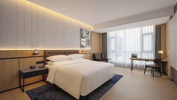 A hotel room with a bed, bedside table, desk, chair, and large windows.