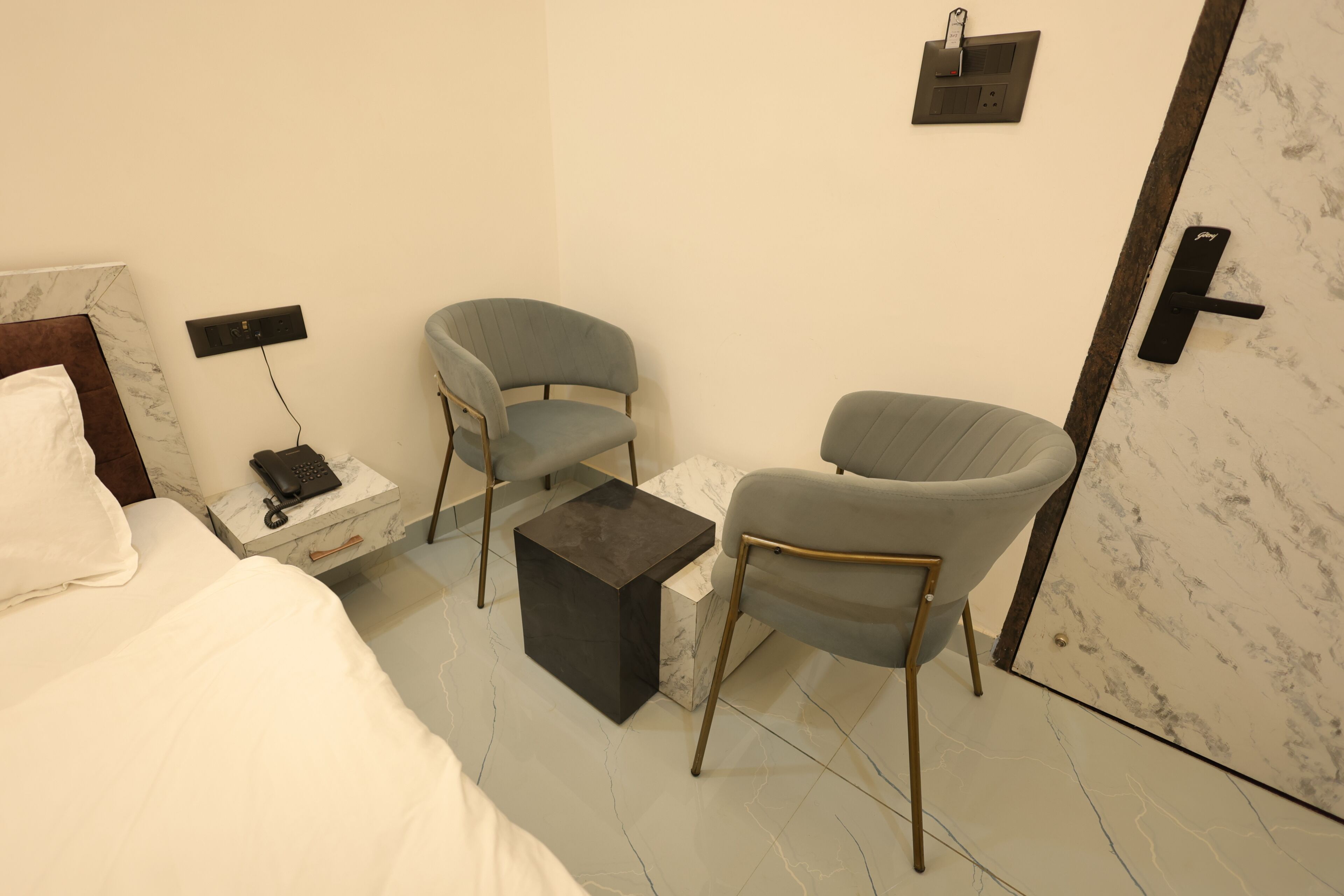 Une chambre d’hôtel comprenant un lit, deux fauteuils, une petite table, une table de chevet avec un téléphone et un interrupteur fixé au mur.