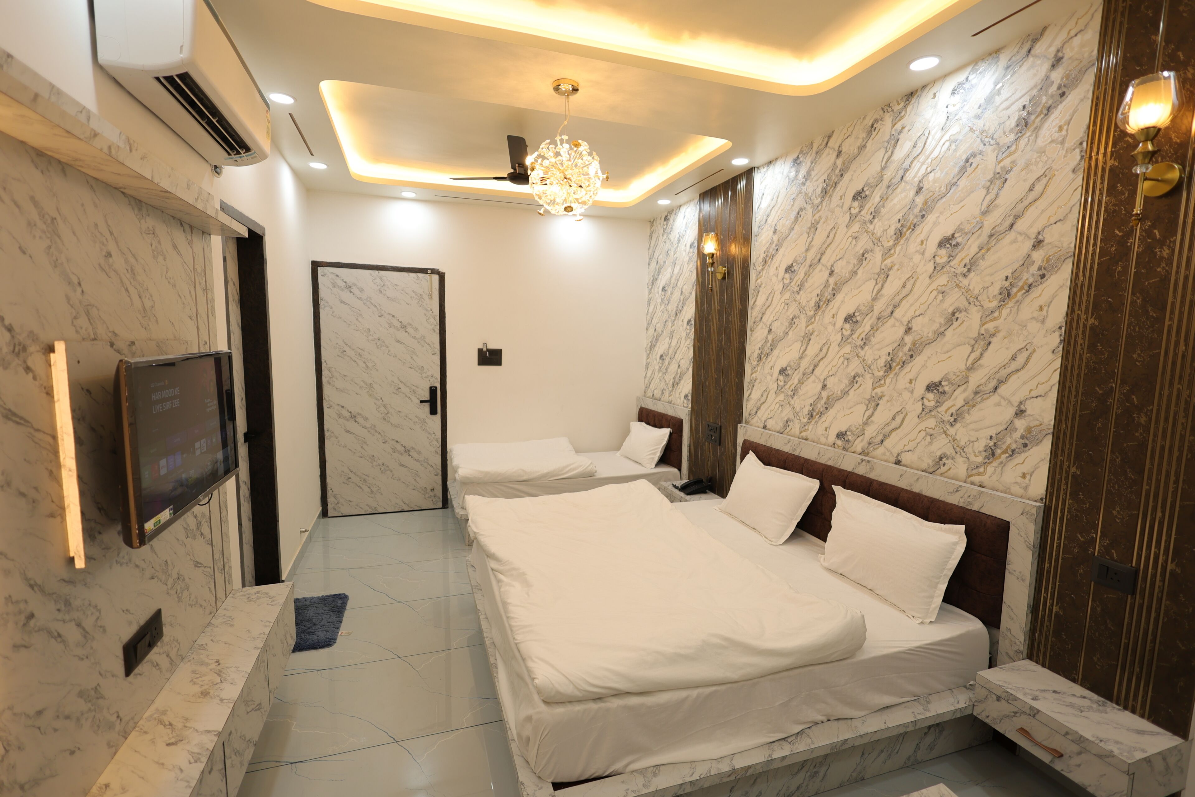 A hotel room with a marble-patterned wall, a large bed with white bedding, a television mounted on the wall, and a ceiling light fixture.