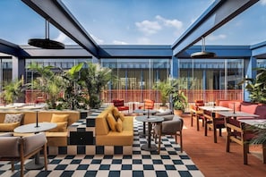 A modern outdoor seating area with checkered flooring, various seating options including sofas and tables, and large windows providing views of the surrounding area.