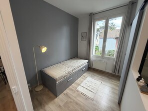 Une piĂšce avec un lit, une lampe sur pied, une fenĂȘtre avec des rideaux et une vue sur les bĂątiments environnants.