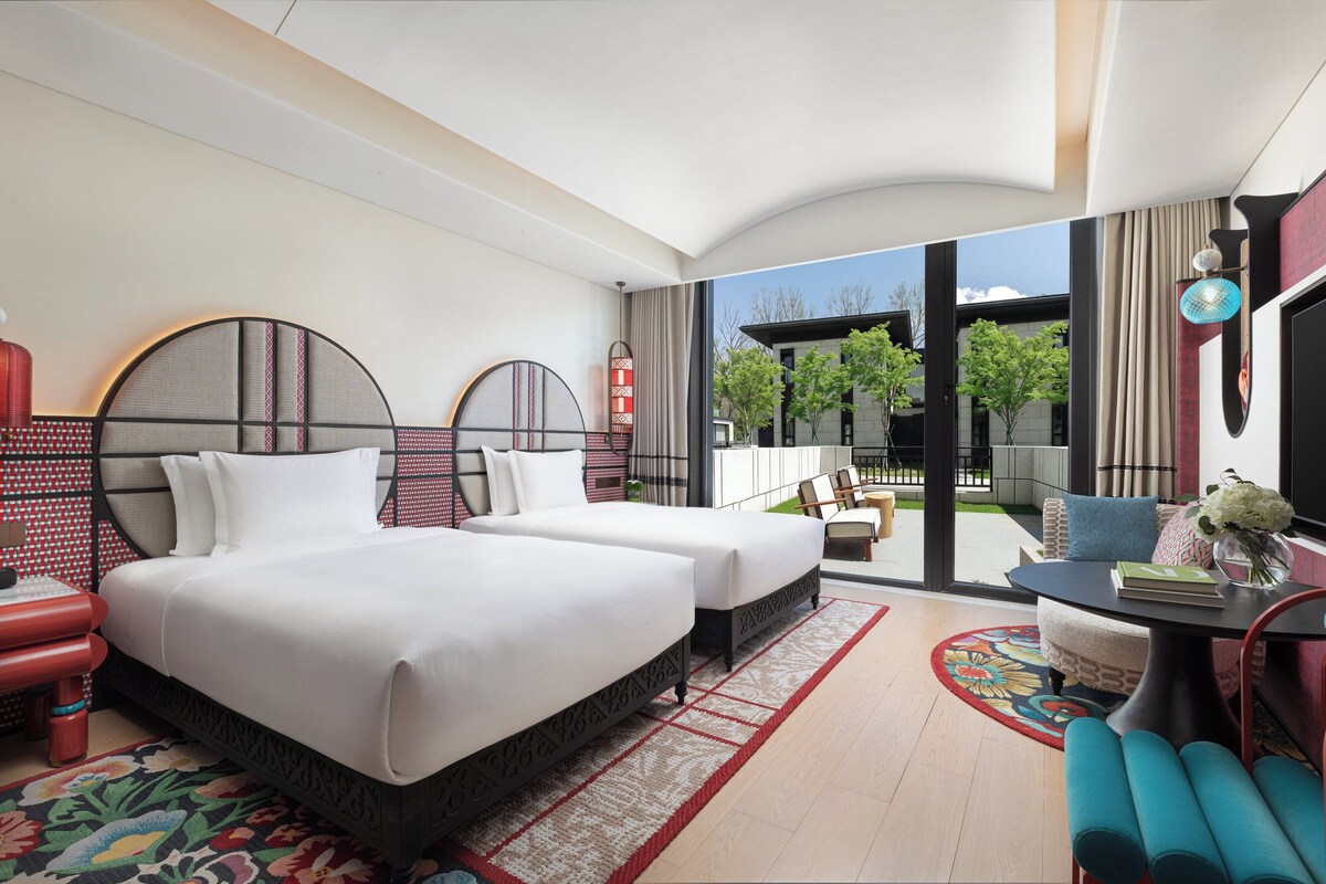 Habitación de hotel con dos camas, una zona de estar con una mesa y sillas, un ventanal con vistas al exterior y un televisor.