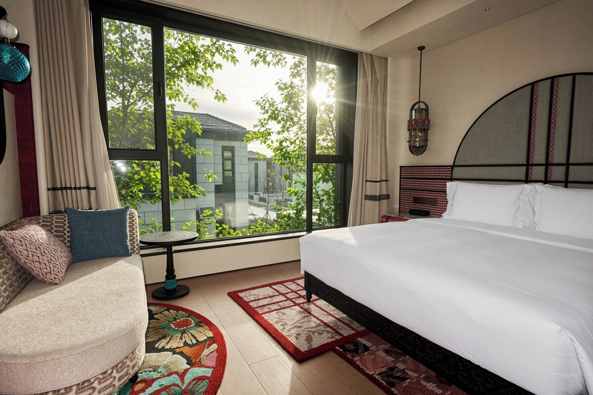 A bedroom with a large bed, a sofa, a small table, and a view of greenery outside.