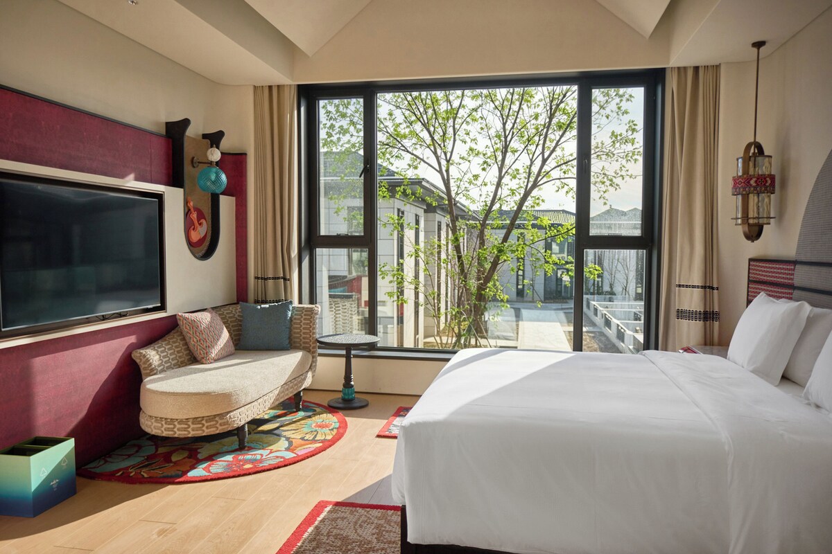 Habitación de hotel con una cama grande, un sofá, un televisor y vistas a un árbol a través de un ventanal amplio.