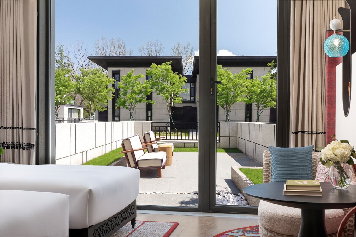 Habitació d'hotel moderna amb una finestra gran, un llit, zona de seients amb cadires, taula petita amb un llibre i flors, i vista a l'exterior de l'edifici.