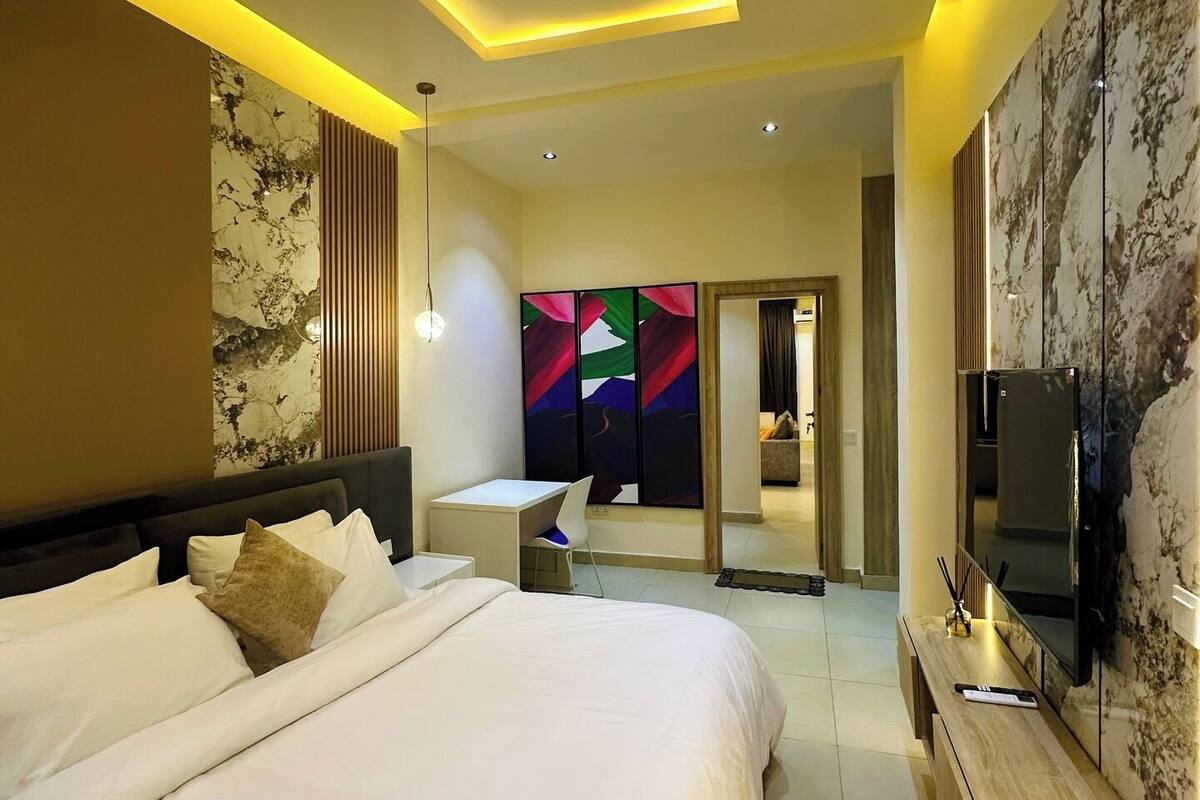A modern hotel room with a large bed, a desk, a TV, and a bathroom visible through a glass door.