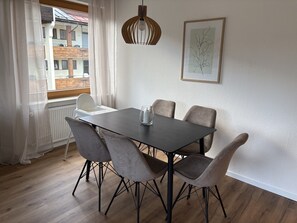 Ein Essbereich mit Tisch und Stühlen, einem Heizkörper, einem Fenster mit Vorhängen und einer gerahmten Pflanze an der Wand.