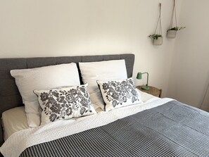 Ein ordentlich bezogenes Bett mit grauem Kopfteil, weißen und gemusterten Kissen, einer karierten Decke und einer grünen Nachttischlampe.