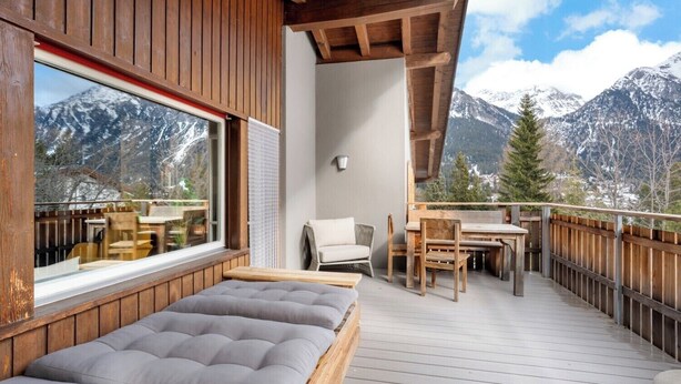 A wooden deck with a bed, a table, and chairs, overlooking snow-capped mountains.