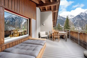 Een houten terras met een bed, een tafel en stoelen, met uitzicht op besneeuwde bergen.