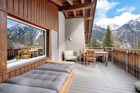 A wooden deck with a bed, a table, and chairs, overlooking snow-capped mountains.