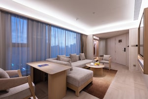 Una habitación de hotel moderna con sofá, una mesa de comedor, una silla, una mesita auxiliar y un ventanal con cortinas.