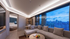 A modern hotel room with a large sofa, a round coffee table with fruit, a bed, and a city view.