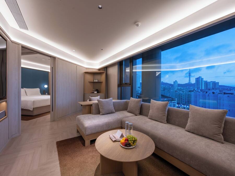 Una habitación de hotel moderna con un sofá grande, una mesa de centro redonda con fruta, una cama y vista a la ciudad.