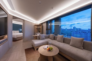 A modern hotel room with a large sofa, a round coffee table with fruit, a bed, and a city view. - Crystal Hotel Qingdao Yan'an 3rd Rd (Qingdao)