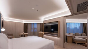 A modern hotel room with a large bed, a TV, a seating area, and a view of the city.