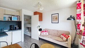 Un espacio habitable compacto con kitchenette, un sofá con cojines estampados, una obra de arte enmarco y una lámpara de pie.