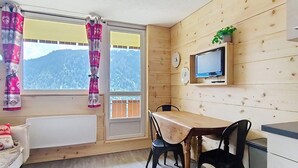 Una habitación con paredes de madera, una mesa con sillas, un televisor montado en la pared y una ventana con cortinas.