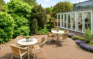 A patio with wicker chairs and a round table, surrounded by lush greenery and a glass conservatory.