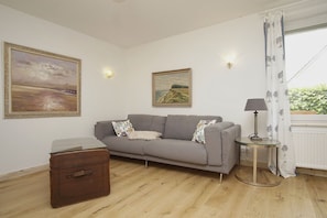 Ein Zimmer mit grauem Sofa, einem Glastisch, einer Lampe, einem Koffer und zwei Bildern an der Wand.