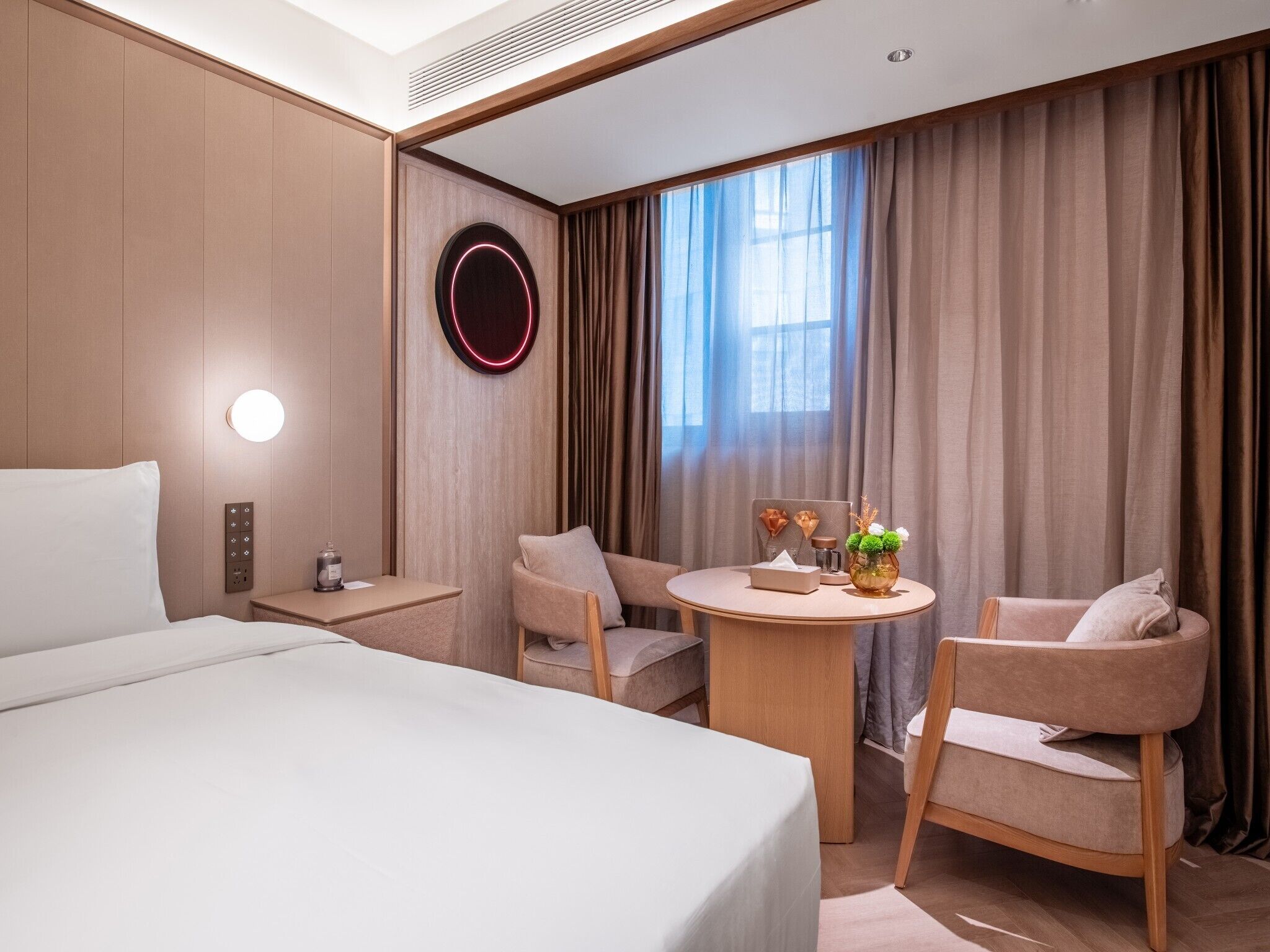 Habitación de hotel con cama, dos sillones, una mesita con un florero y un adorno redondo en la pared.