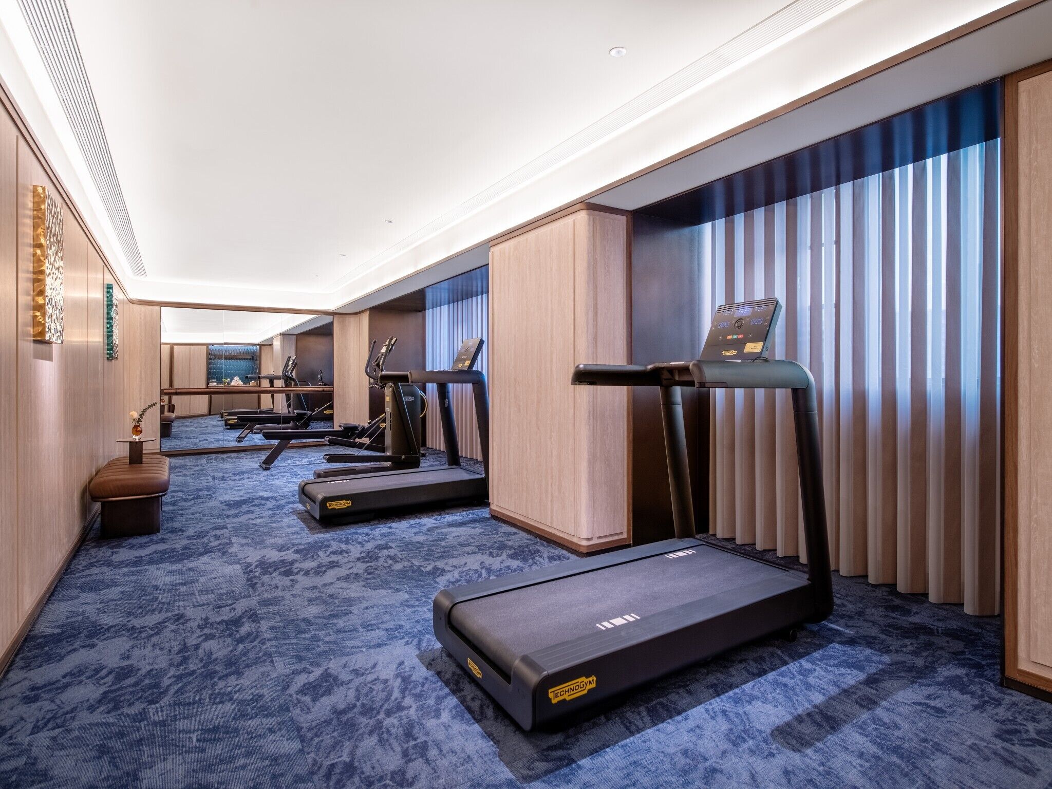 Una camera d'hotel con area fitness che include tapis roulant e attrezzature per esercizi, un'ampia finestra con persiane e pavimento in moquette.