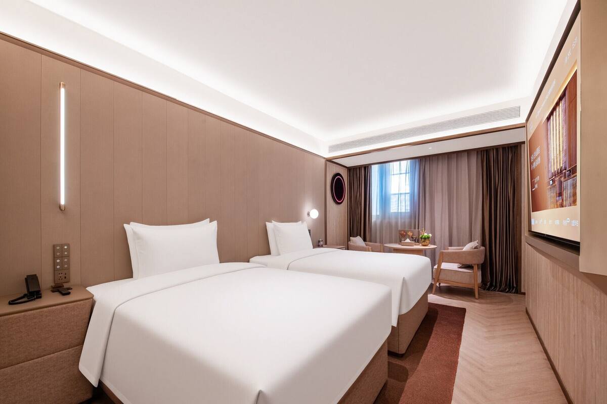 A hotel room with two beds, a desk, a chair, a TV, and a large window with curtains.