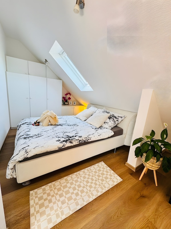 Une chambre à coucher avec un lit, une lucarne, une table de chevet avec une lampe, une plante en pot et un plancher en bois.