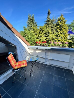 A balcony with a glass table and a plaid chair, surrounded by greenery and a blue building. - Apartment "Adlerhorst" with Wi-Fi and Air Conditioning (Worpswede)