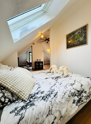 A bedroom with a skylight, a bed with a patterned bedspread, a painting on the wall, and a ceiling fan. - Apartment "Adlerhorst" with Wi-Fi and Air Conditioning (Worpswede)