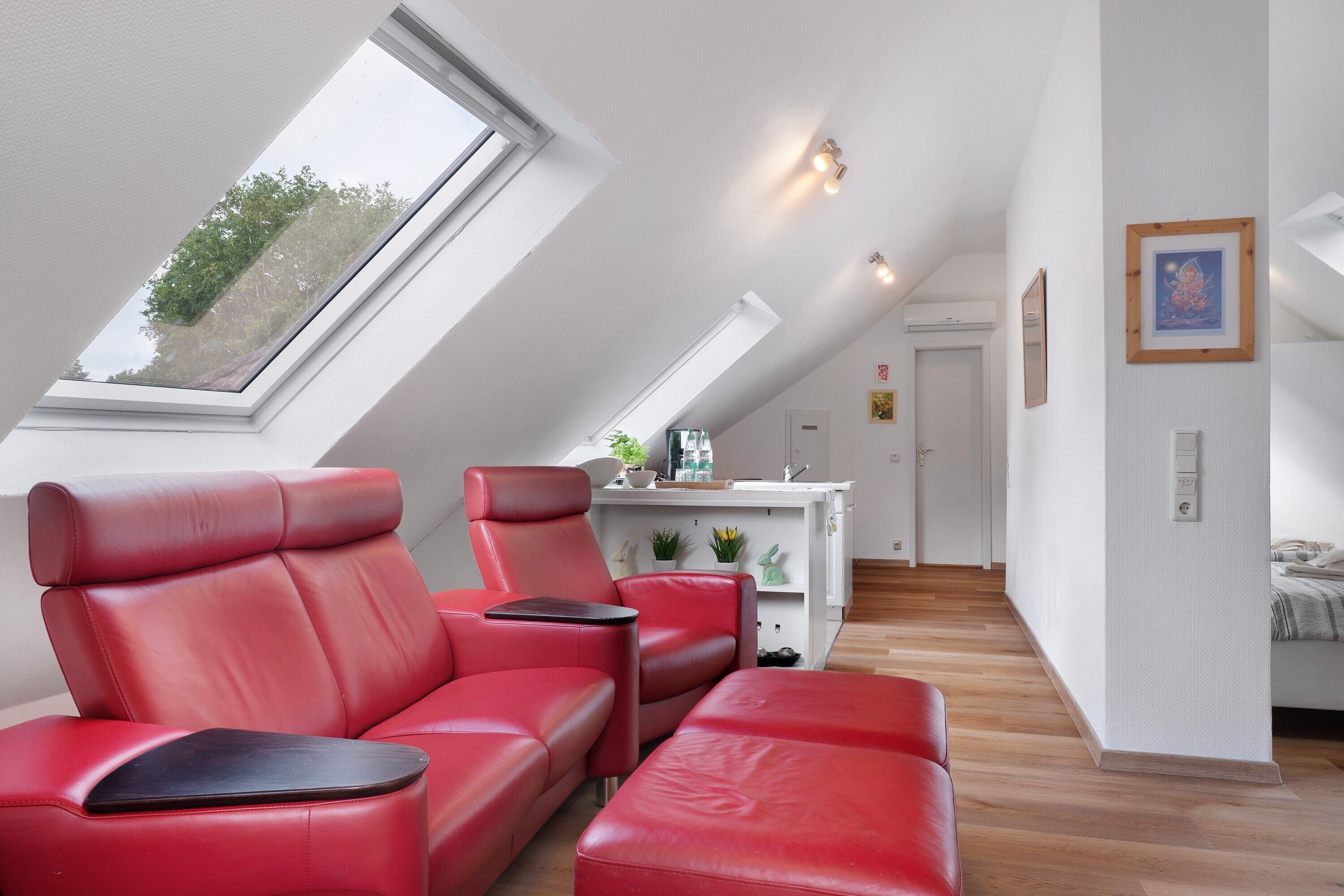 Une chambre de style loft moderne, avec un plafond en pente, un coin salon en cuir rouge, une petite kitchenette et une vue sur le ciel grâce à une lucarne.