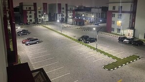 Un estacionamiento con varios autos, un edificio de fachada roja y blanca, y una entrada bien iluminada.