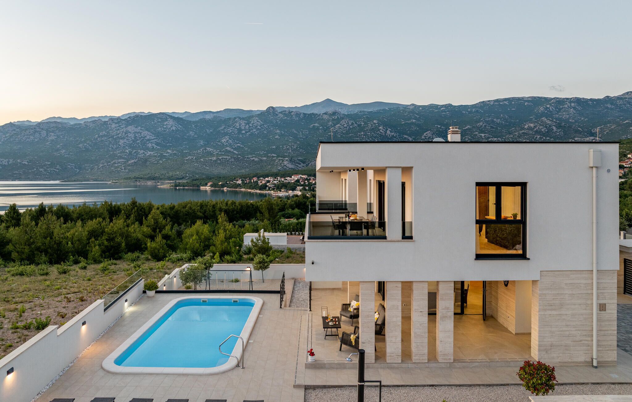 A modern two-story house with a swimming pool, a balcony, and a view of a lake and mountains.