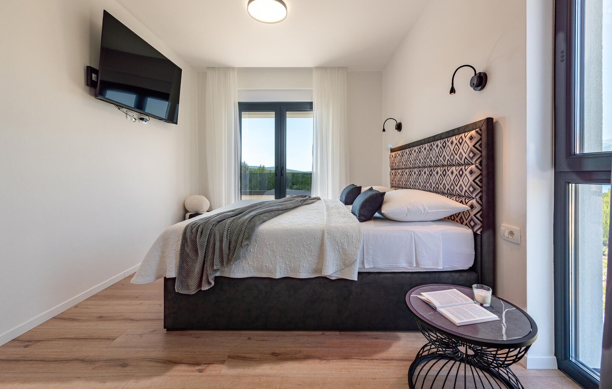 Een slaapkamer met een bed, een nachtkastje met een boek en een glas, een aan de muur bevestigde televisie en een raam met uitzicht.