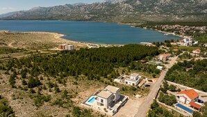Uma área costeira com uma vasta extensão de água, montanhas ao fundo e uma zona residencial com casas e piscina.