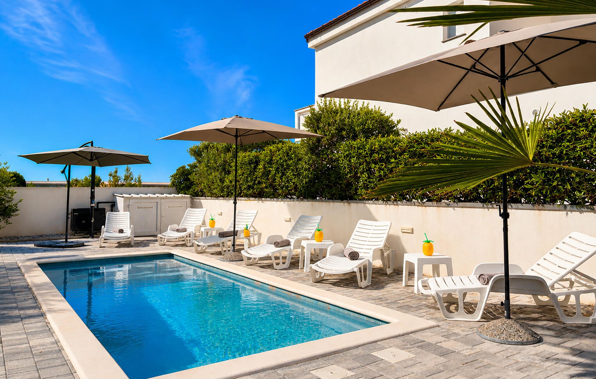 Une piscine entourée d’un bâtiment blanc et de verdure, avec des chaises longues et des parasols à proximité.