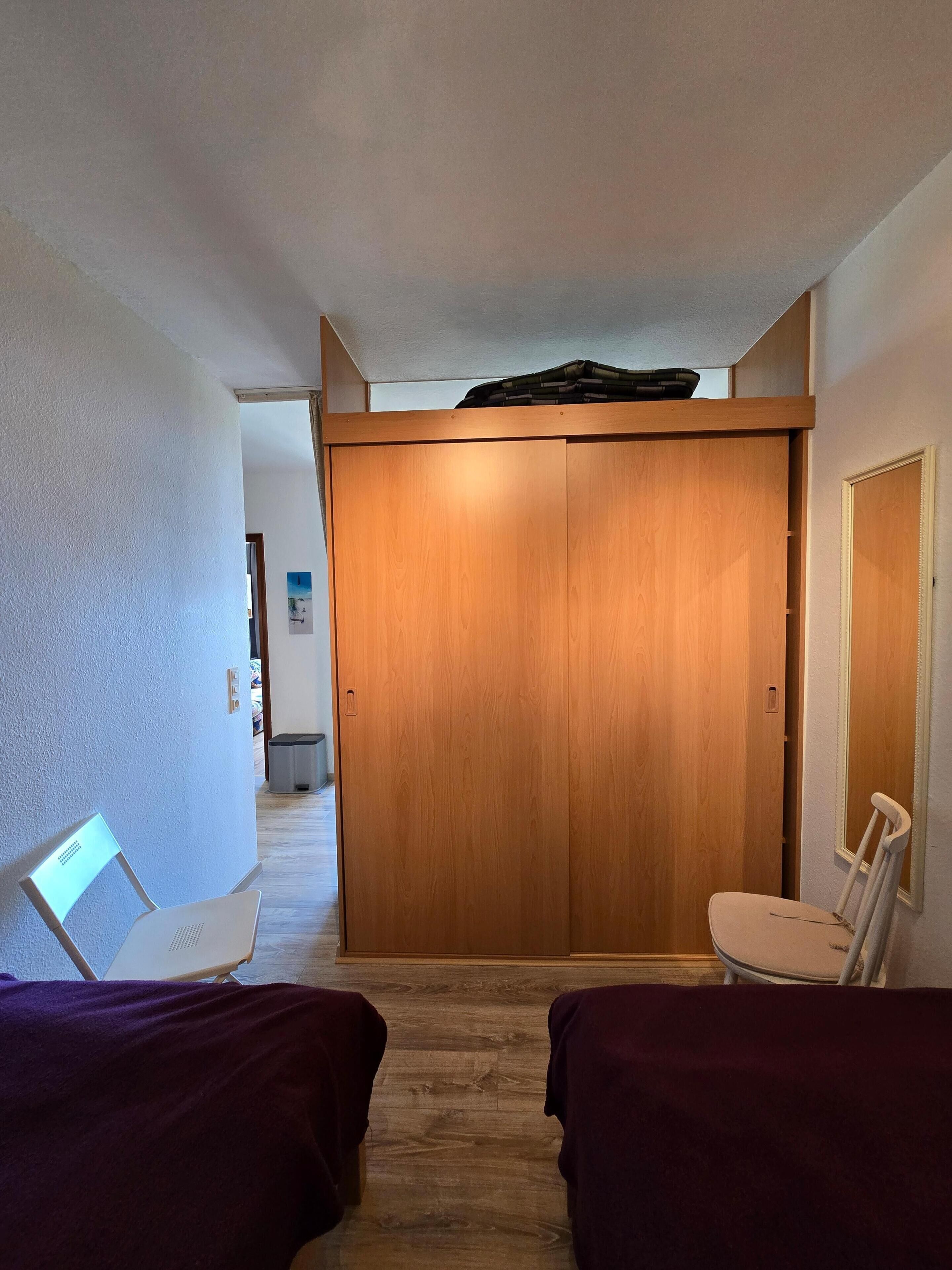 Ein Zimmer mit Holzschrank, einem Sessel, einem Schreibtisch mit Laptop und einem Bett.
