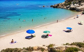 Ein Strand mit klarem türkisblauem Wasser, darin schwimmende Menschen und Sonnenschirme im Sand.