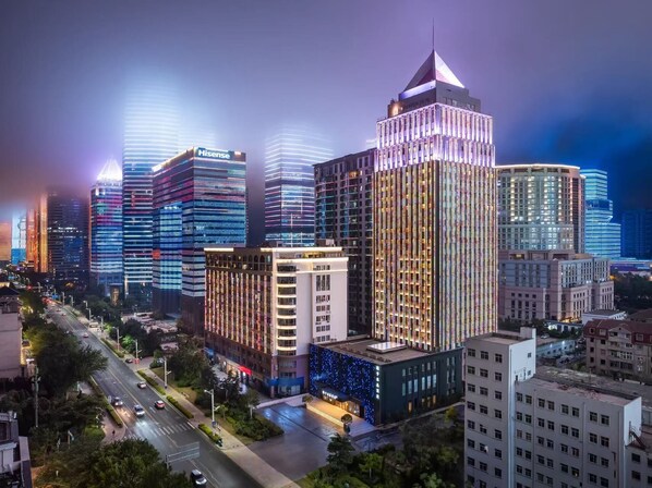 Paisagem urbana com edifícios altos modernos, um hotel imponente com topo triangular característico e uma rua bem iluminada.