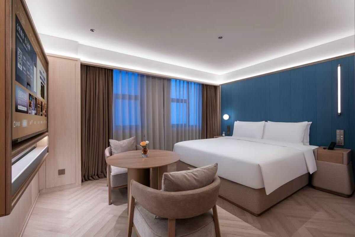 Ein Hotelzimmer mit einem großen Bett, einem Schreibtisch mit Stuhl, einem kleinen Tisch mit Vase und einem an der Wand befestigten Fernseher.