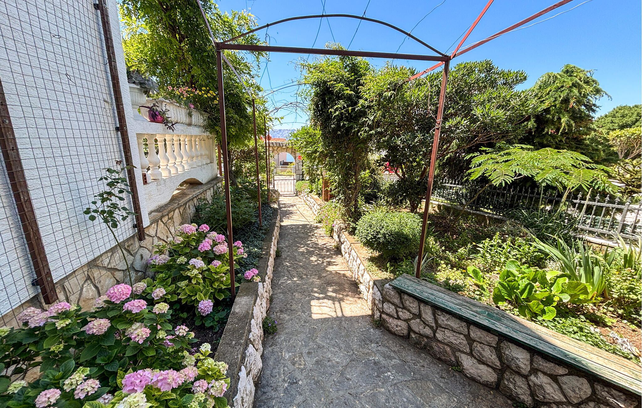 Un étroit sentier de jardin, bordé d’un côté par un mur de pierre et de l’autre par une clôture en métal, qui mène à un portail.