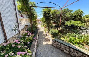Ein schmaler Gartenweg, an einer Seite von einer Steinmauer und an der anderen von einem Metallzaun gesäumt, der zu einem Tor führt.