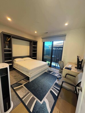 Une chambre à coucher avec un lit, un bureau avec un ordinateur, une chaise, un radiateur et une vue sur l’extérieur.