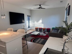 Ein modernes Wohnzimmer mit Flachbildfernseher, Deckenventilator, Essbereich mit Tisch und Stühlen sowie einer Couch mit farbenfrohen Kissen.