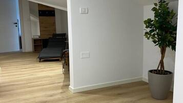 A room with a wooden floor, a potted plant, and a bed in the background.