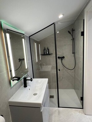 Une salle de bain moderne dotée d’une douche à parois de verre, d’un lavabo blanc avec un robinet noir et d’un miroir avec éclairage intégré.