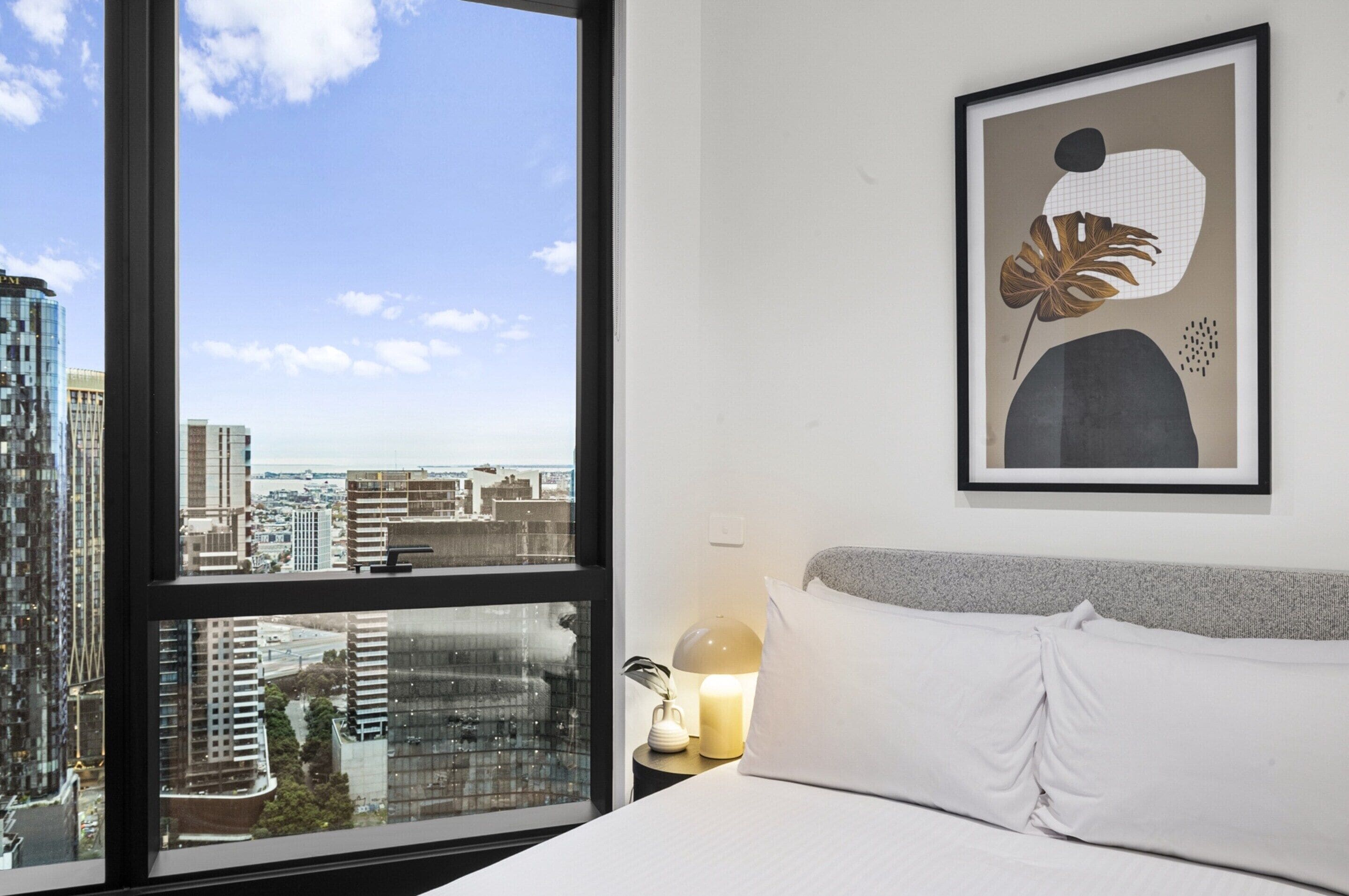 Ein Hotelzimmer mit großem Fenster, einem Bett, einem Nachttisch mit Lampe und einem gerahmten Kunstwerk an der Wand.