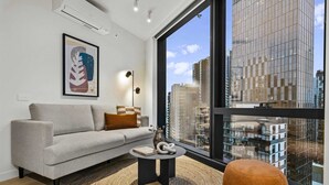 A modern living room with a sofa, a round coffee table, a large window with a city view, and a framed artwork on the wall. - High-rise 2-bed Stay at Queensbridge (Melbourne)