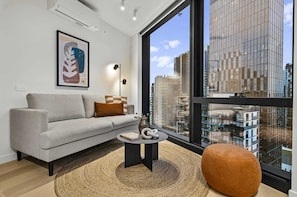 Ein modernes Wohnzimmer mit einer Couch, einem runden Couchtisch, einem großen Fenster mit Stadtblick und einem gerahmten Kunstwerk an der Wand.