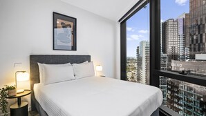 A modern hotel room with a large bed, two bedside tables with lamps, a framed artwork above the bed, and a city view through large windows. - High-rise 2-bed Stay at Queensbridge (Melbourne)